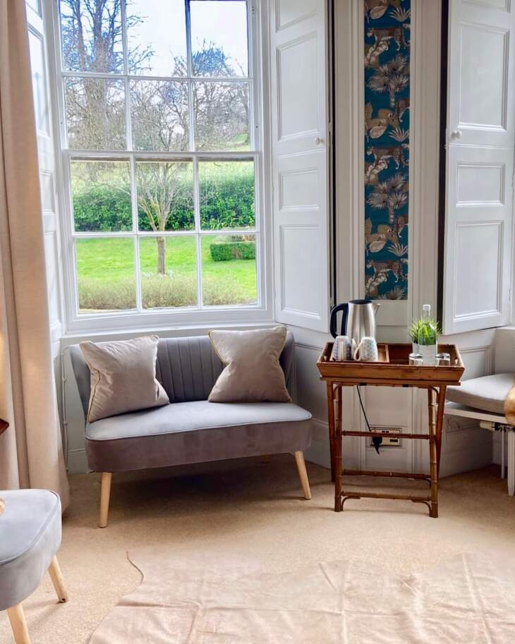 Cozy hotel sitting area with velvet chairs, fur throws, patterned wallpaper, large windows showcasing green garden views, and a tea tray.