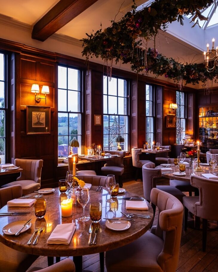 Elegant dining room with plush chairs, candlelit tables, large windows showing scenic countryside, and festive holiday decor with chandelier