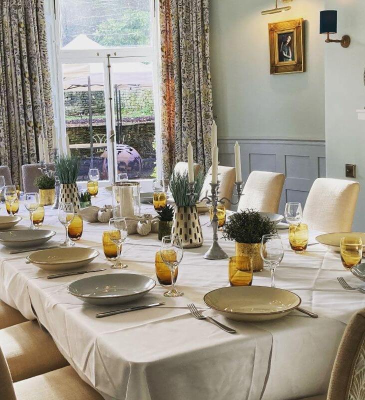 Elegant dining room with a long table set for 12, white linens, amber glasses, candelabras, and floral curtains by large windows.