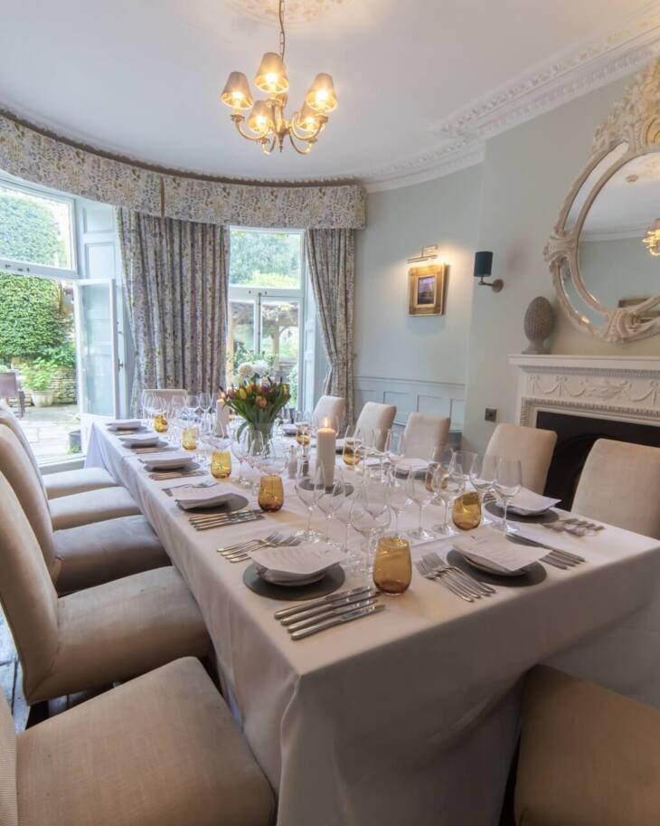 Elegant dining room with ornate fireplace, large round mirror, beige upholstered chairs, floral curtains, and table set for a formal meal
