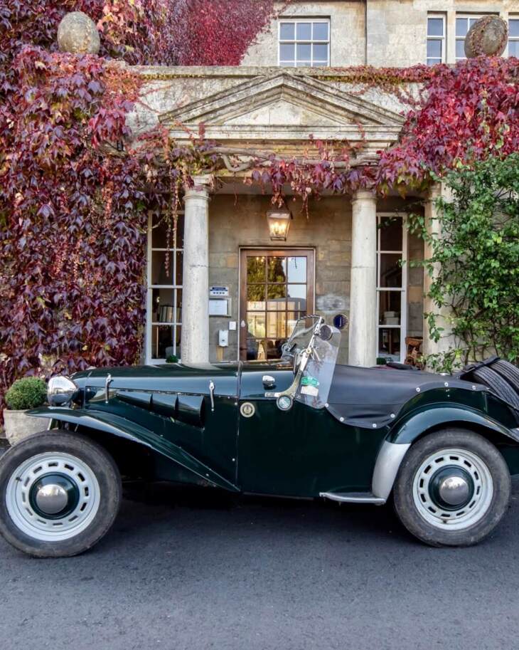 Classic vintage convertible parked in front of a historic stone hotel entrance covered in red ivy with tall columns and large windows