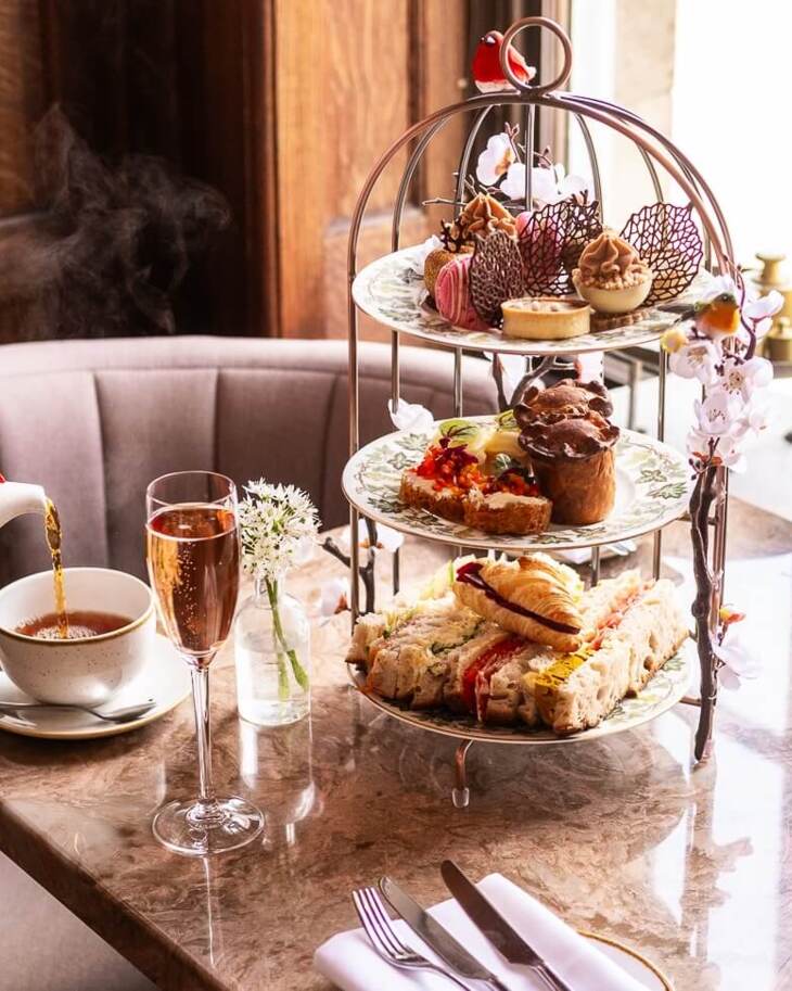 Afternoon tea setup with tiered platter of sandwiches, pastries, and desserts, steaming tea poured into a cup, and two glasses of sparkling wine