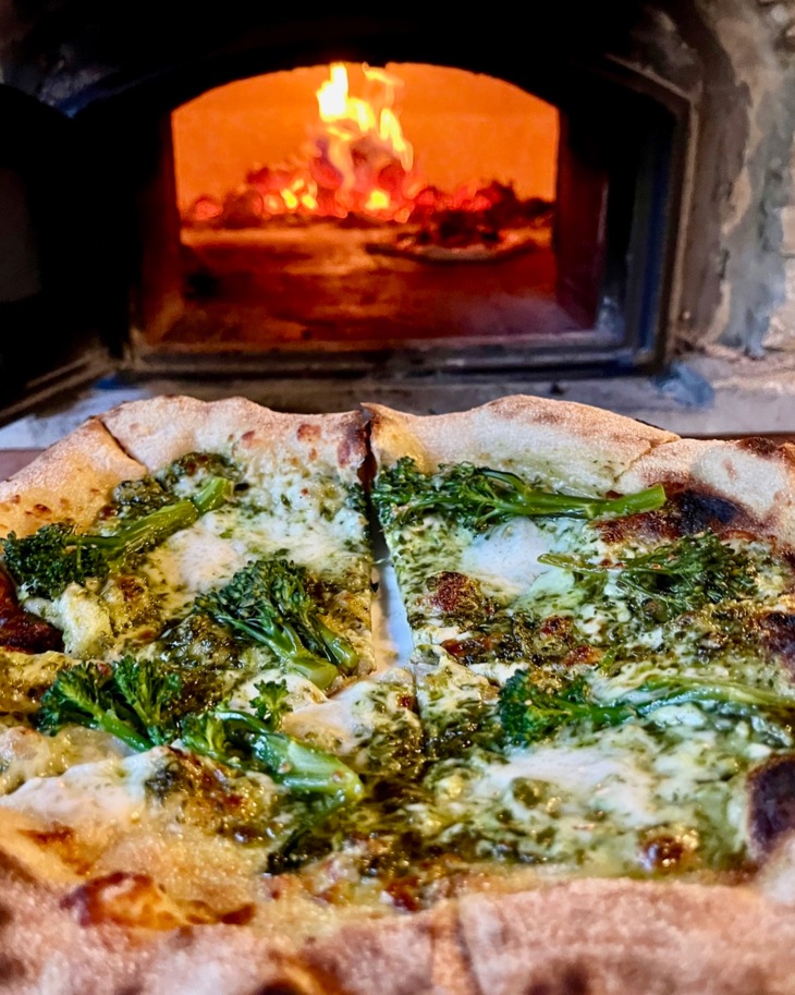 Wood-fired pizza topped with melted cheese and steamed broccoli in front of a rustic stone pizza oven