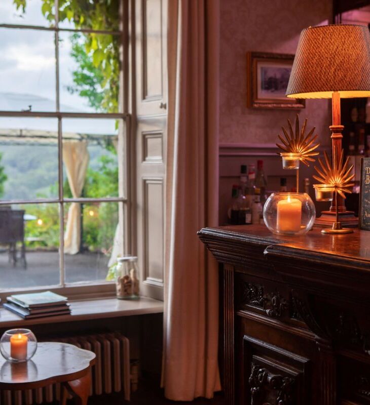 Cozy hotel bar with antique wooden counter, glowing candlelight, decorative lamps, and large window overlooking outdoor seating and greenery