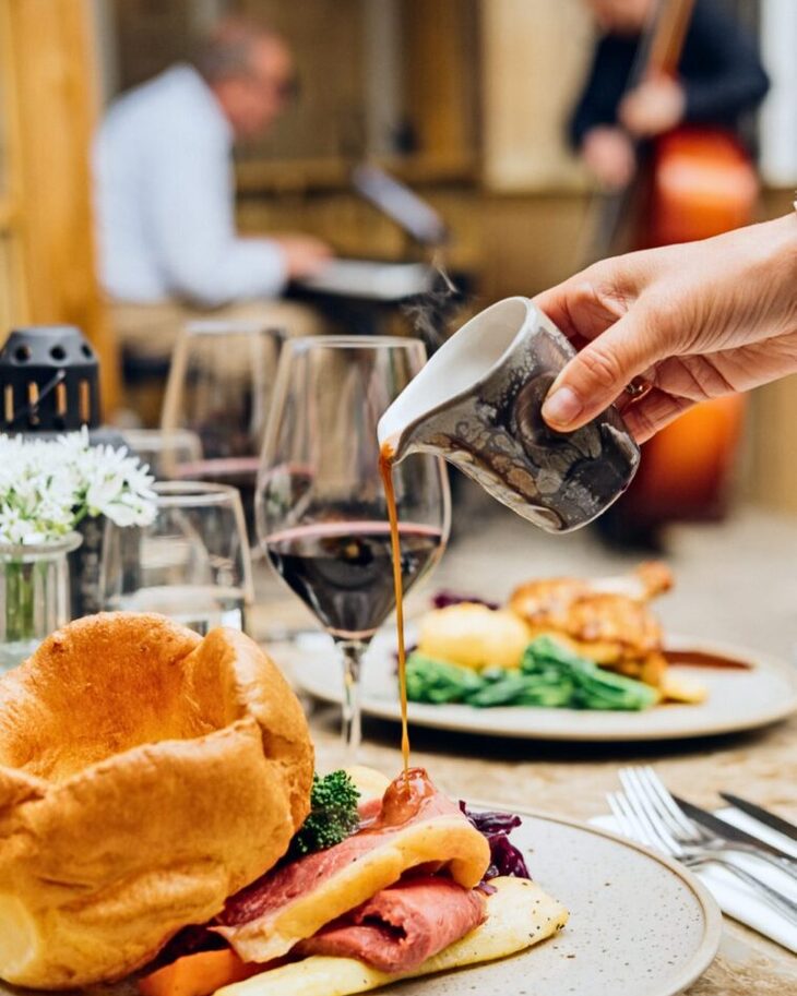 Hand pouring gravy over classic roast beef with Yorkshire pudding, vegetables, and wine glasses in an elegant dining setting