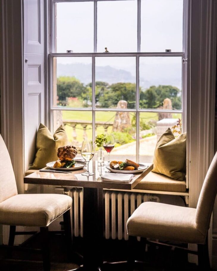 Cozy dining nook with cushioned window seats, two chairs, plates of gourmet food, wine glasses, overlooking lush garden and countryside.