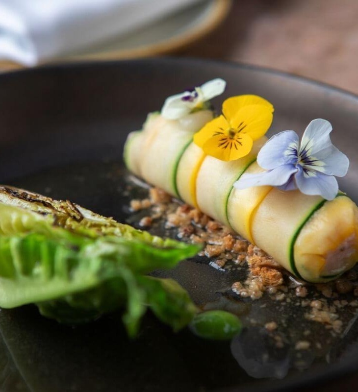 Gourmet dish with rolled vegetable wrapped in thin slices, topped with edible flowers, served with grilled lettuce on black plate