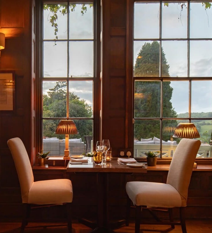 Cozy dining table for two by tall windows with soft lamps, overlooking lush greenery and outdoor seating at sunset.