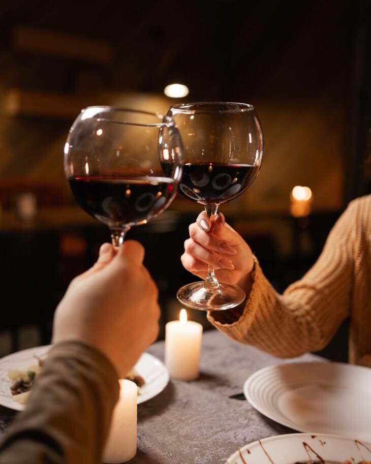 Two guests toasting with red wine glasses at a candlelit table with gourmet dishes in a cozy restaurant ambiance