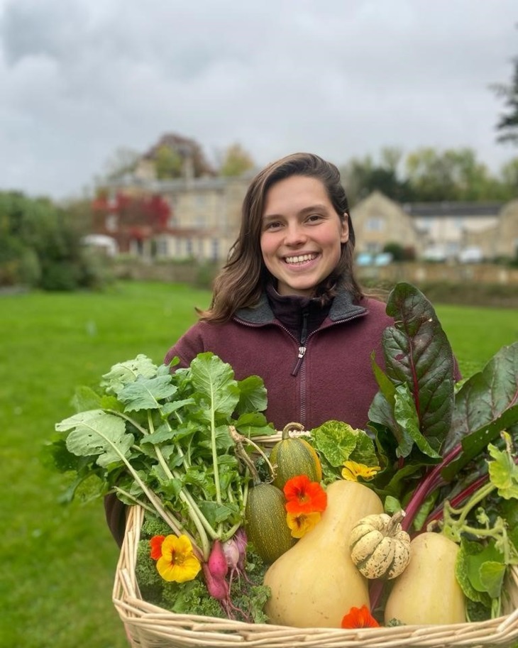 Smiling woman holds basket of fresh garden vegetables and vibrant flowers in outdoor hotel garden area