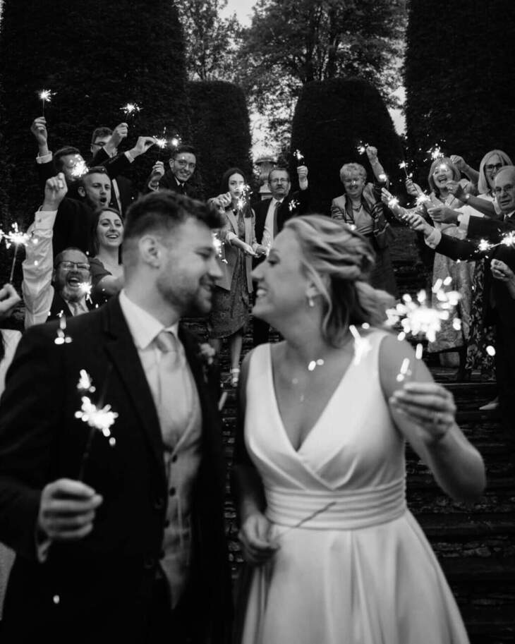 Newlywed couple smiling and holding sparklers while guests celebrate with sparklers on outdoor stone steps at wedding reception