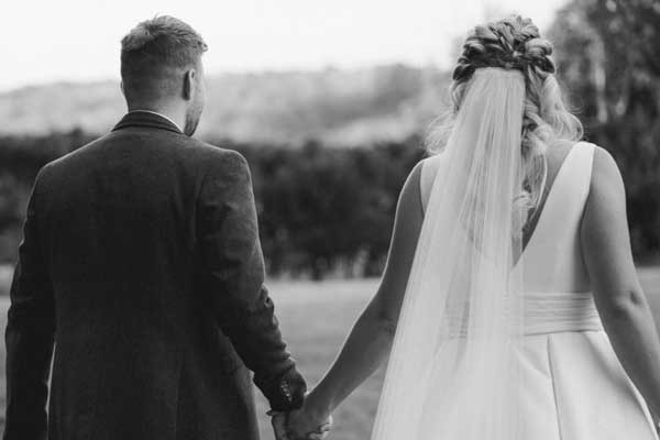 Newlywed couple holding hands walking outdoors in wedding attire, featuring a bridal gown with long veil and groom in suit jacket