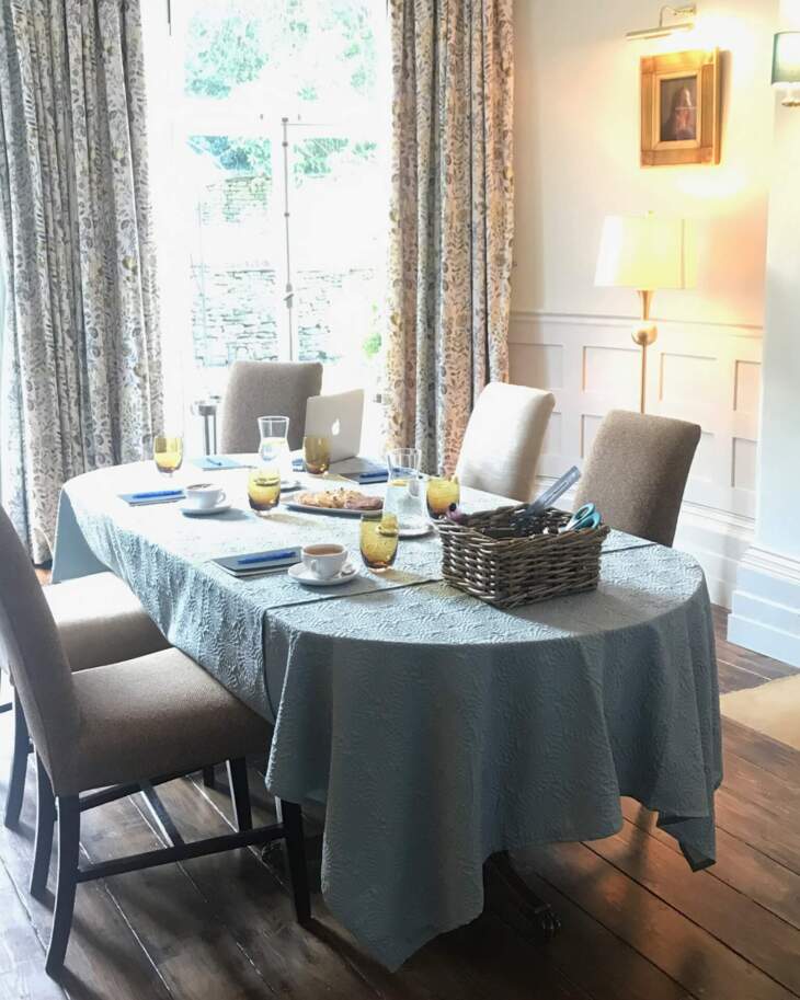 Bright dining room with a long table covered in a textured blue tablecloth, surrounded by cushioned chairs and set with glasses and cups
