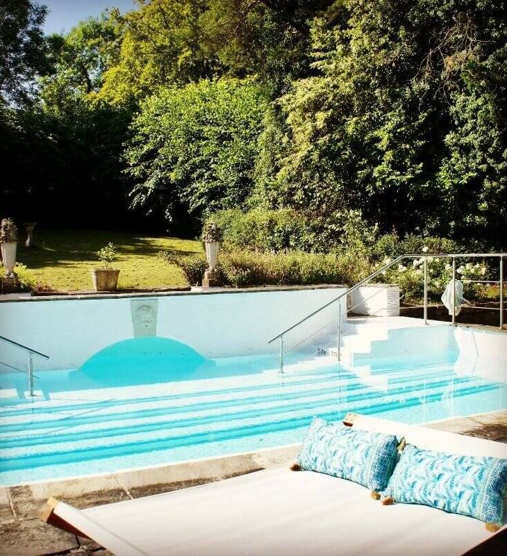 Outdoor hotel pool with calm blue water, wide steps, handrails, surrounded by lush green trees and a cushioned lounge bed nearby
