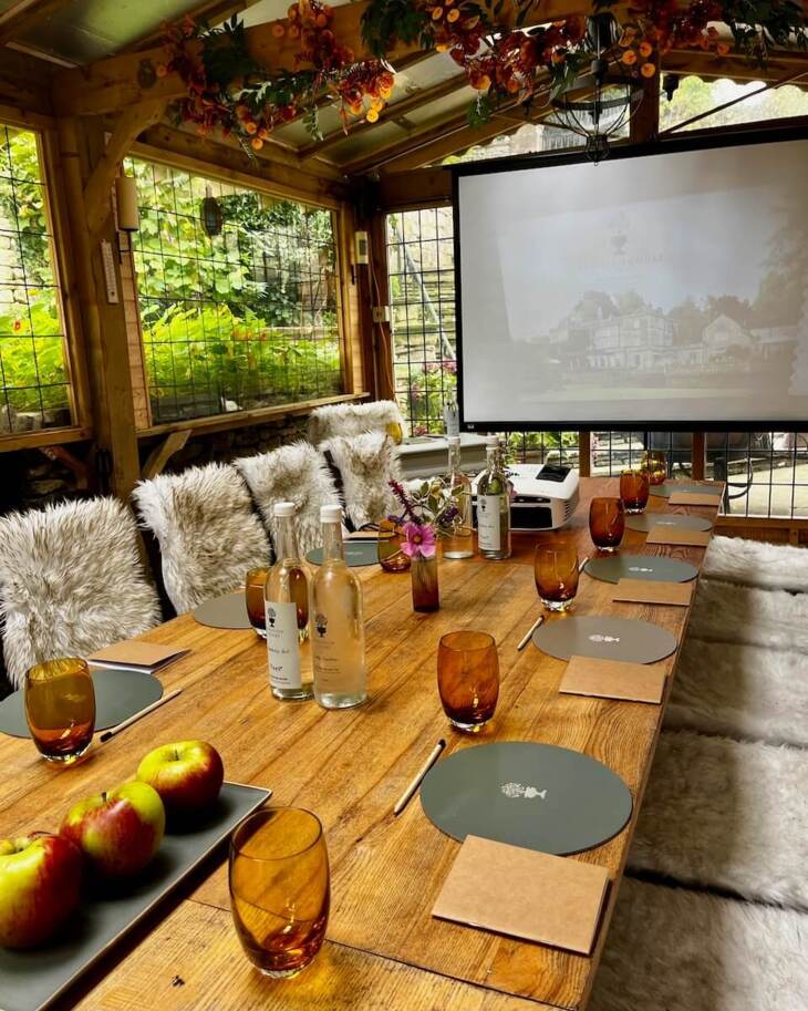 Cozy wooden meeting room with furry chair covers, amber glasses, apples, bottled water, notepads, and a projector screen in garden setting