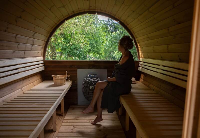 Guest wrapped in towel relaxing inside a wooden barrel sauna with large arched window overlooking lush green trees