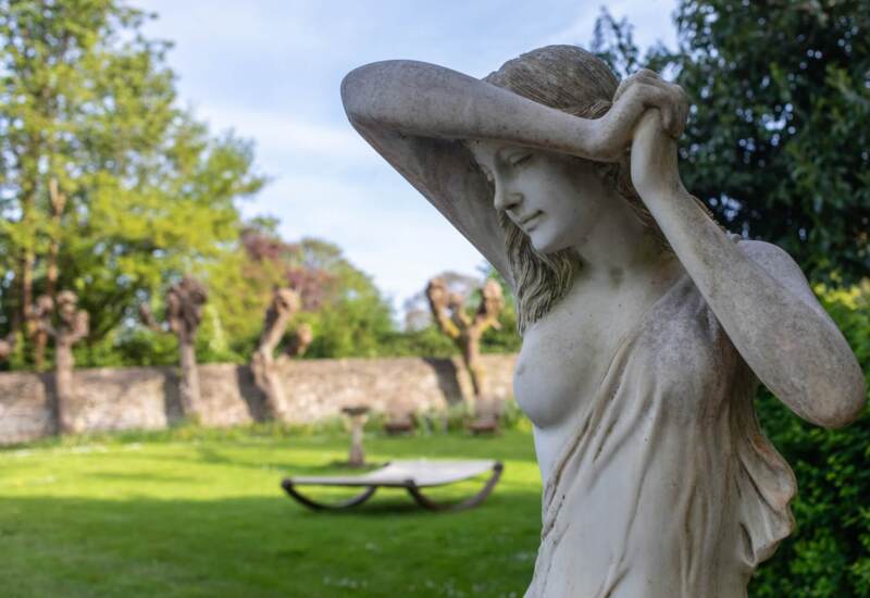Elegant stone statue of a woman in a lush garden with trimmed trees and stone wall in the background
