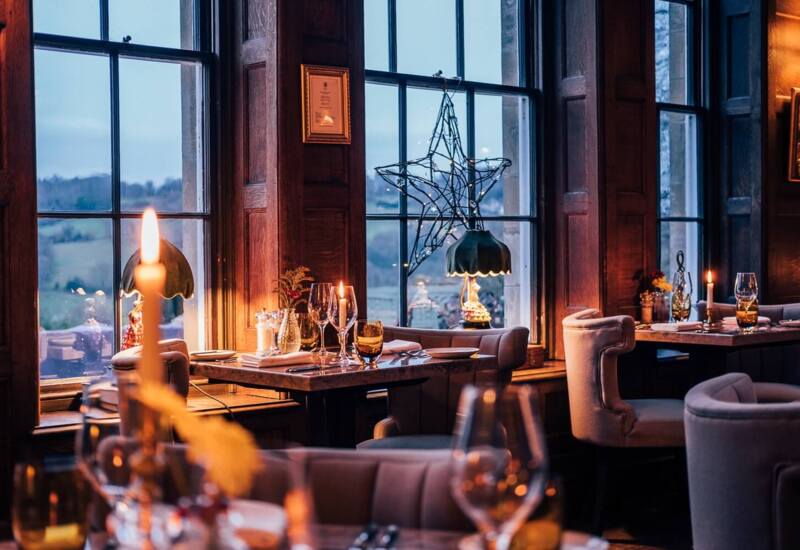 Cozy fine dining room with wood paneling, candlelit tables, plush chairs, decorative lamps, and large windows overlooking a countryside view