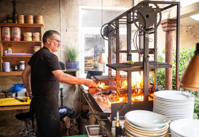 Chef grilling food over an open flame in a rustic kitchen area with shelves of ingredients and stacked white plates nearby