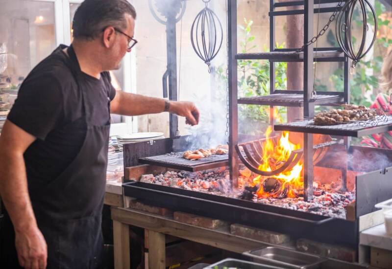 Chef grilling skewers over an open flame grill with smoke rising in a rustic outdoor kitchen setting