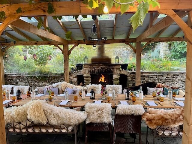 Cozy outdoor dining area with wooden tables, sheepskin-covered chairs, and a stone fireplace under a wooden pergola surrounded by greenery.