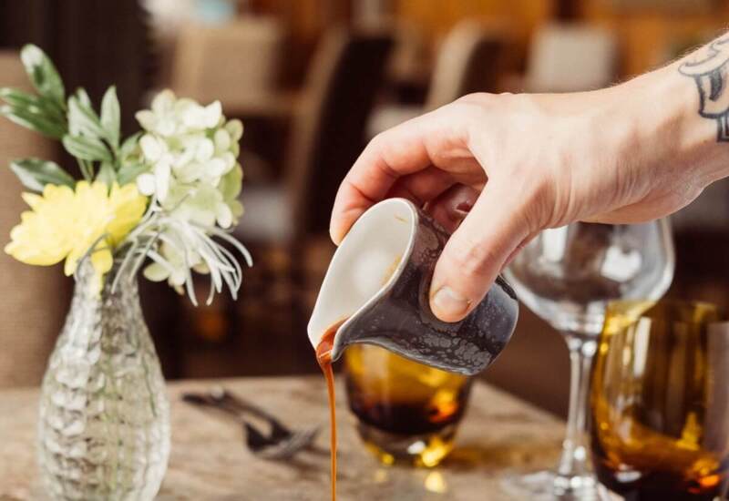 Hand pouring sauce over a plated gourmet pork dish with roasted vegetables in an elegant dining setting with glassware and fresh flowers