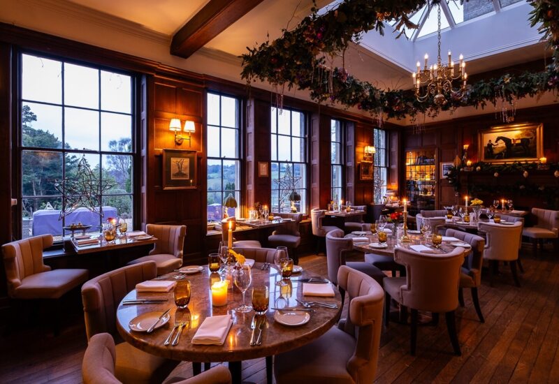 Elegant dining room with plush chairs, candlelit tables, large windows showing scenic countryside, and festive holiday decor with chandelier
