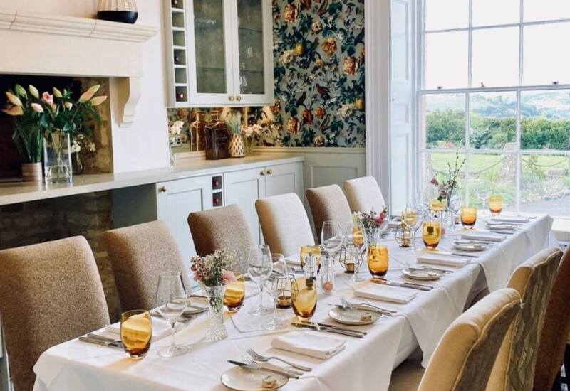 Elegant dining room with long table set for guests, amber glassware, floral centerpieces, cozy chairs, and large window overlooking green landscape