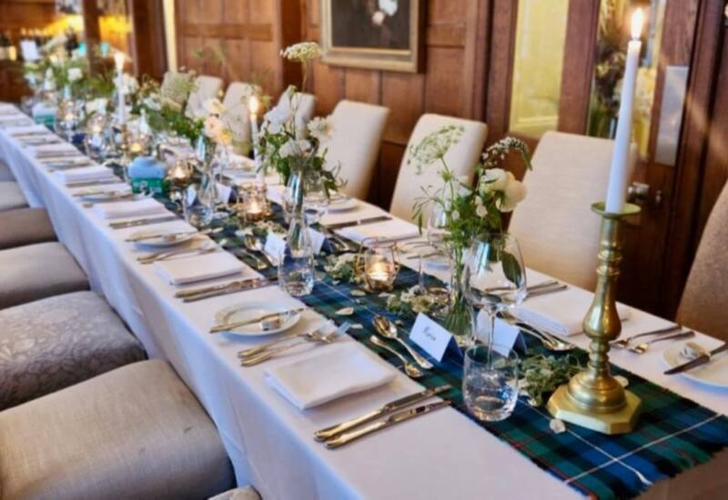 Elegant dining table set with white linens, glassware, fresh floral arrangements, and brass candlesticks in a wood-paneled room