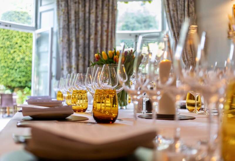 Elegant dining table set with folded napkins, amber glassware, wine glasses, candles, and a floral centerpiece near large windows