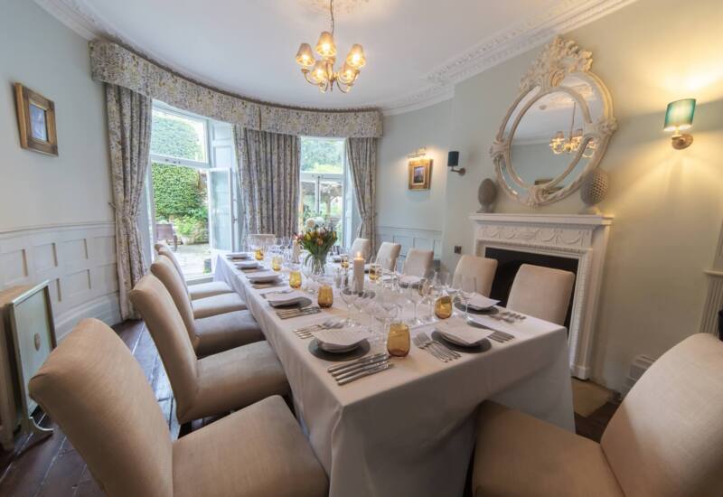 Elegant dining room with ornate fireplace, large round mirror, beige upholstered chairs, floral curtains, and table set for a formal meal