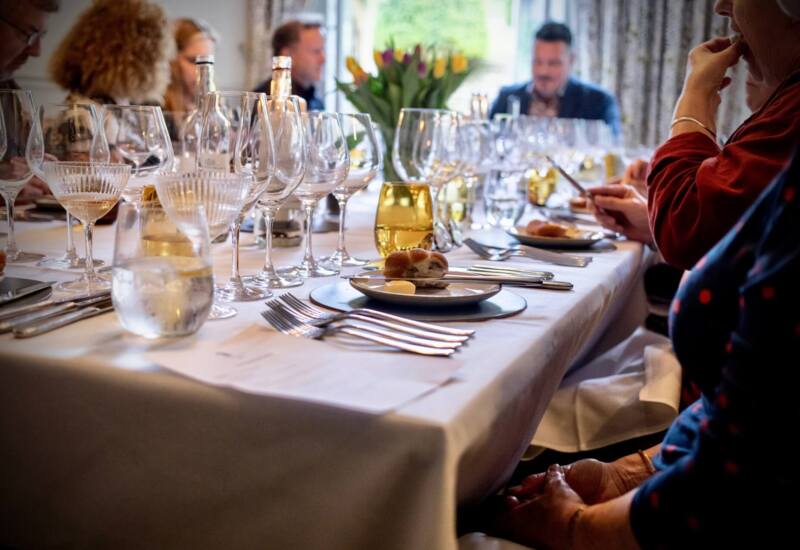 Elegant dining table set with multiple wine glasses, bread rolls, and butter, guests engaged in conversation in a cozy restaurant.