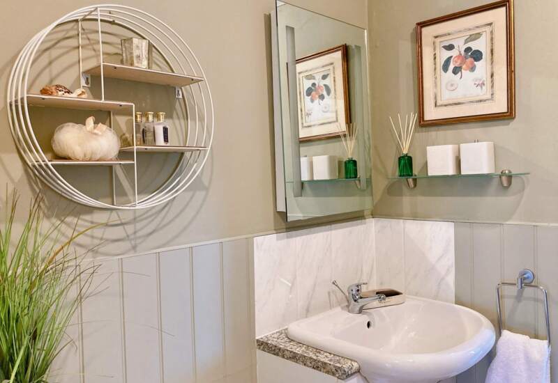 Bright bathroom with white sink, granite countertop, round wall shelf with toiletries, floral artwork, and a hand towel