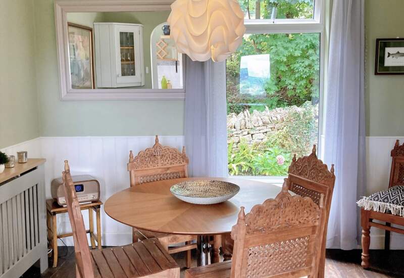 Cozy dining area with round wooden table, carved wooden chairs, soft green walls, decorative pendant light, and garden view through window