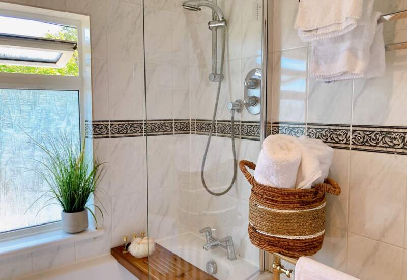 Bright hotel bathroom with marble tiles, walk-in shower, glass partition, rolled white towels in woven basket, and decorative plant by frosted window