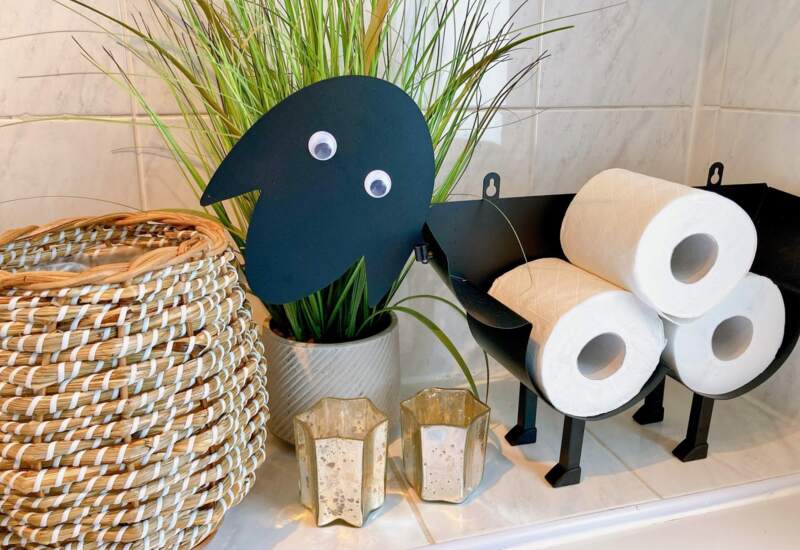 Bathroom shelf with rolled white toilet paper in black holder shaped like a sheep, green plant in textured pot, two small candle holders, and woven basket