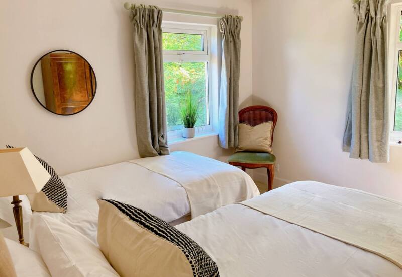 Bright twin bedroom featuring white bedding, patterned pillows, green cushioned chair, round wall mirror, and windows with beige curtains overlooking greenery