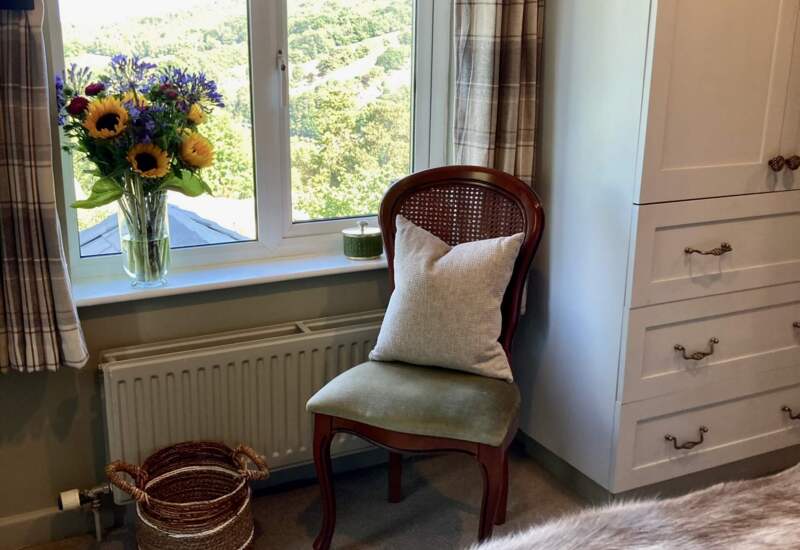 Cozy hotel room corner with cushioned wooden chair, plaid curtains, fresh flowers by window, and soft fur throw on bed.