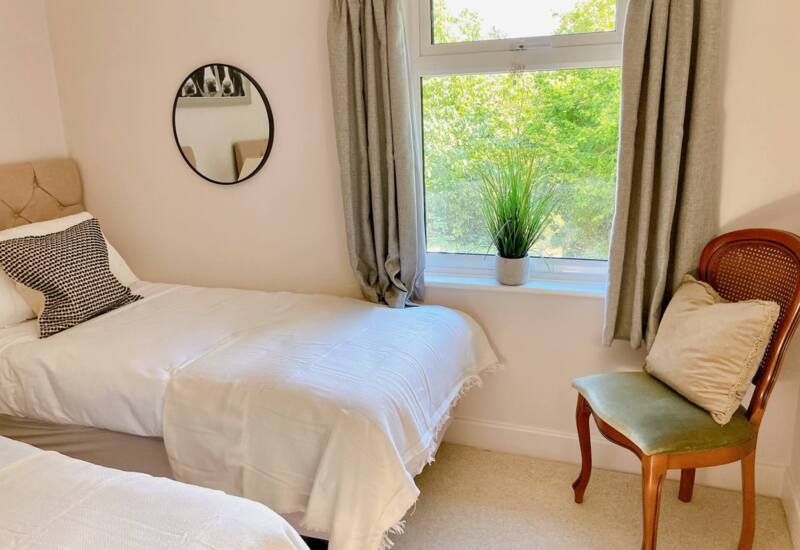 Cozy twin bed room with neutral decor, round wall mirror, window overlooking greenery, and wooden chair with cushion