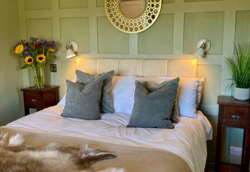 Cozy bedroom with a soft beige bedspread, gray pillows, fur throw, paneled green walls, floral vase, and decorative round mirror