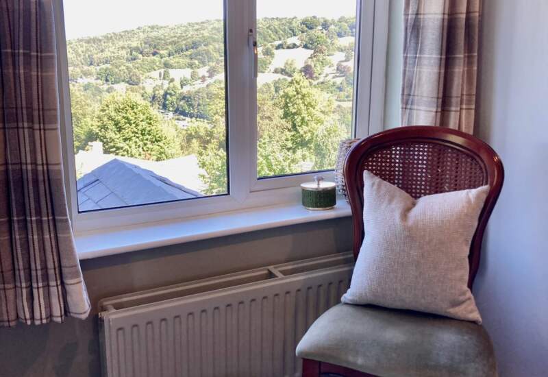 Cozy corner with cushioned wooden chair, woven basket, window with plaid curtains, and scenic view of green hills outside