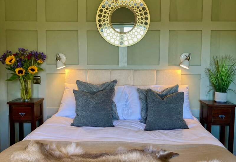 Cozy hotel bedroom with paneled green wall, white and gray bedding, fur throw, bedside lamps, fresh flowers, and potted plant