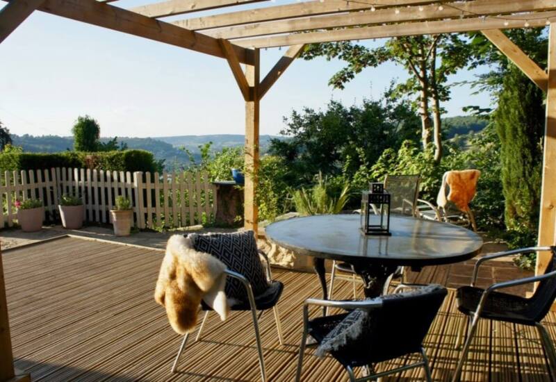 Sunny wooden terrace with round table, metal chairs draped with cozy throws, overlooking lush green hills and garden plants