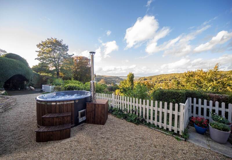 Outdoor bubbling hot tub with wooden steps beside a white picket fence overlooking lush green hills under a blue sky