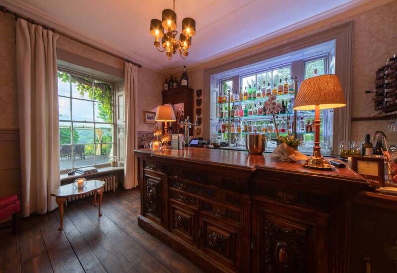 Cozy hotel bar with ornate wooden counter, shelves of liquor bottles by large windows overlooking a green outdoor terrace