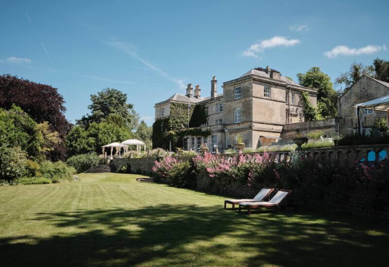 Historic stone hotel surrounded by lush gardens, pink flowers, and sun loungers on a manicured lawn under a clear blue sky