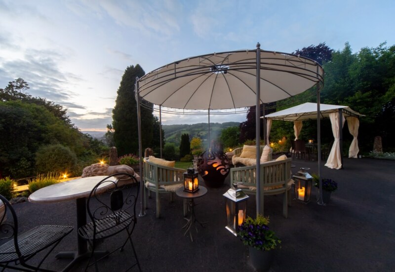 Outdoor seating area with cushioned benches under canopy, lanterns, fire pit, and scenic garden views at dusk