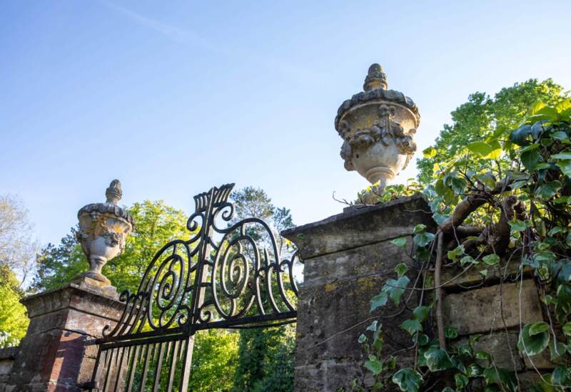 Ornate wrought iron gate with detailed stone urns and lush greenery in a serene garden setting at a historic hotel estate