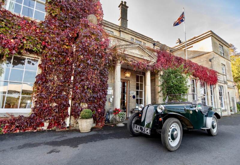Historic stone hotel entrance with red ivy, classic convertible car parked outside, and British flag fluttering above the building
