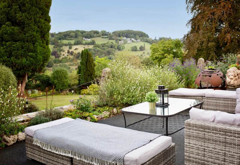 Outdoor patio with cushioned wicker seating, glass coffee table, decorative lantern, and scenic hillside views surrounded by lush greenery